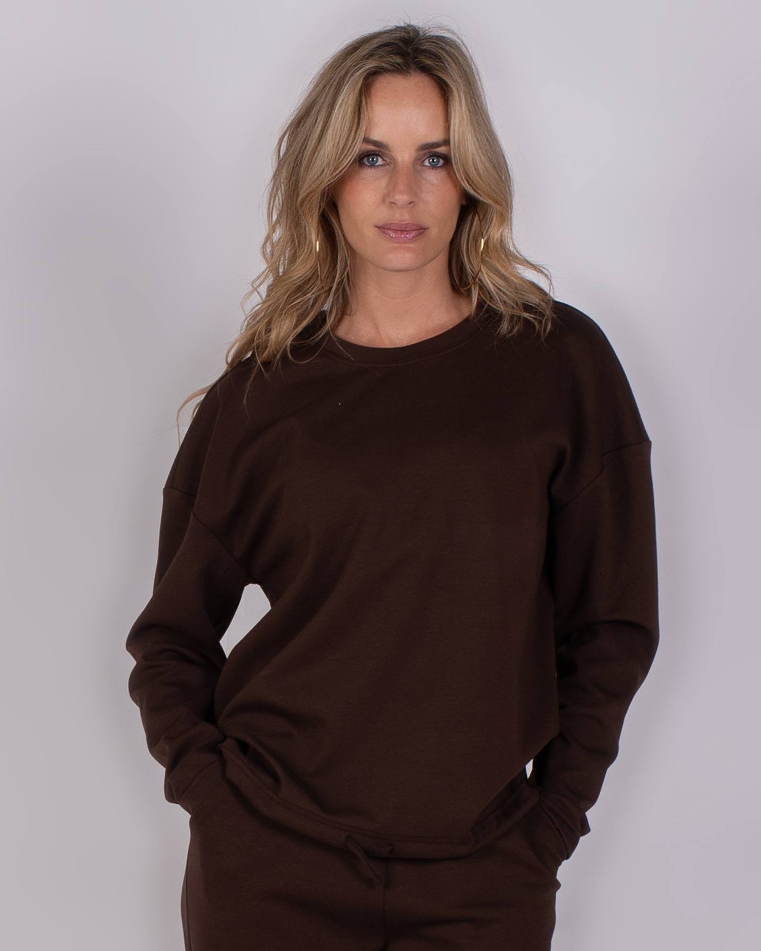 Vrouw poseert in een donkerbruine sweater en bijpassende broek uit de Studio AW25-26 collectie.