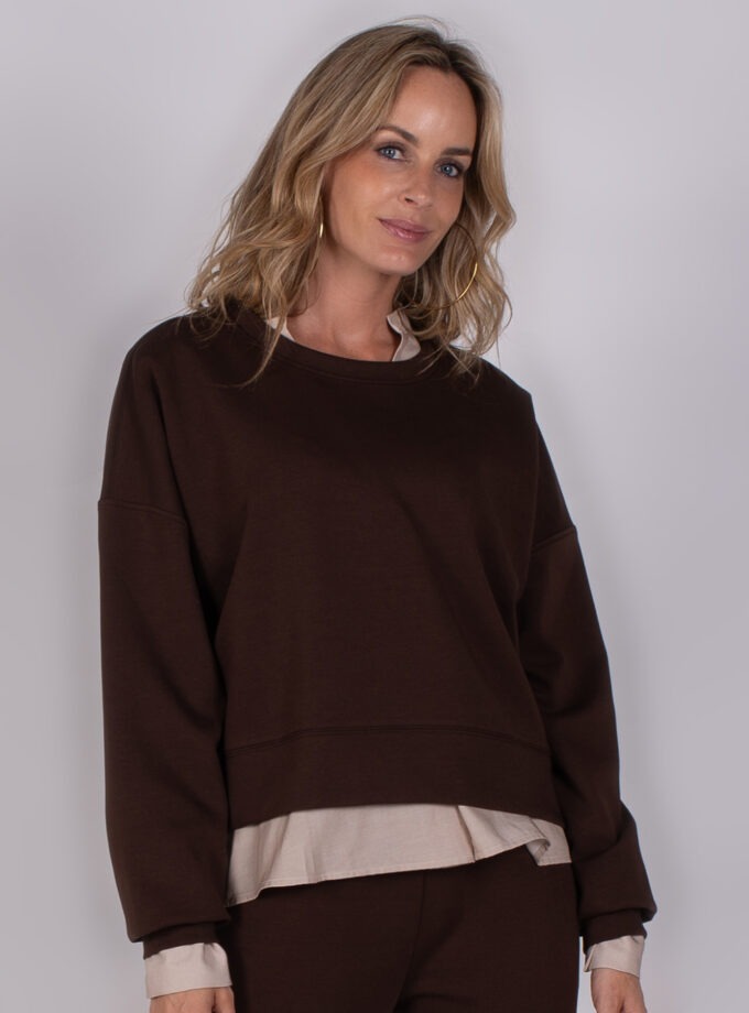 Vrouw draagt een donkerbruine oversized sweater met beige blouse eronder uit de Studio AW25-26 collectie.