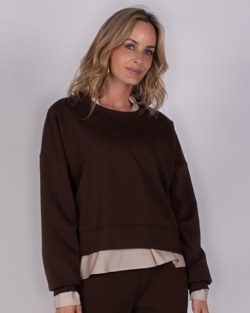 Vrouw draagt een donkerbruine oversized sweater met beige blouse eronder uit de Studio AW25-26 collectie.