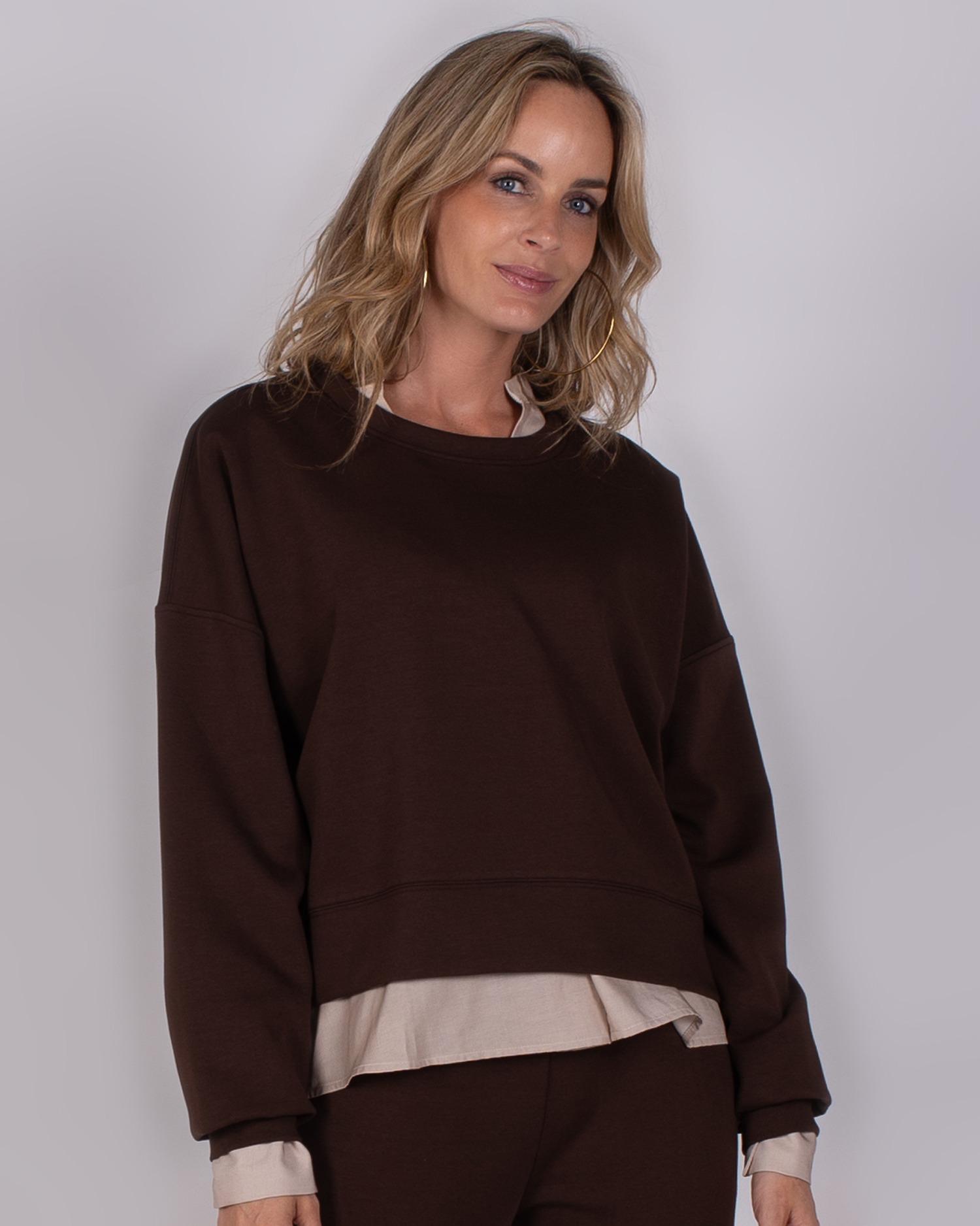 Vrouw draagt een donkerbruine oversized sweater met beige blouse eronder uit de Studio AW25-26 collectie.