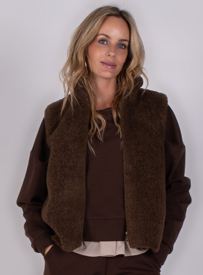 Vrouw met losse blonde haren draagt een donkerbruine teddy bodywarmer over een bijpassende bruine trui.