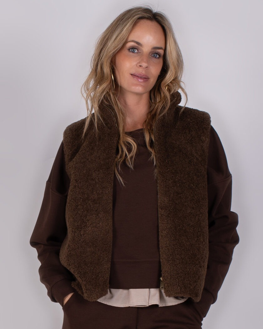 Vrouw met losse blonde haren draagt een donkerbruine teddy bodywarmer over een bijpassende bruine trui.