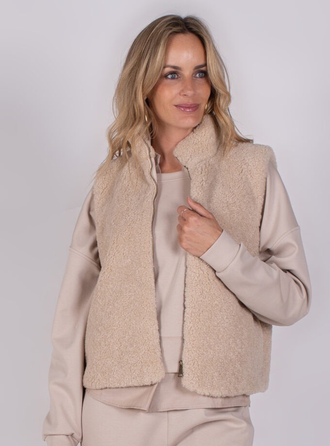 Vrouw draagt een beige teddy bodywarmer over een lichtbeige loungepak tegen een neutrale achtergrond.