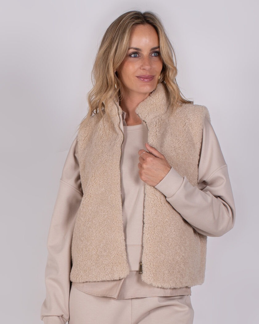 Vrouw draagt een beige teddy bodywarmer over een lichtbeige loungepak tegen een neutrale achtergrond.