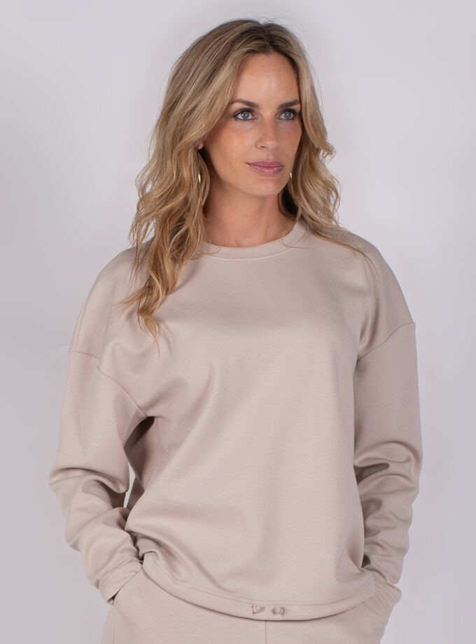 Vrouw draagt een beige trui uit de Studio AW25-26 collectie tegen een neutrale achtergrond.