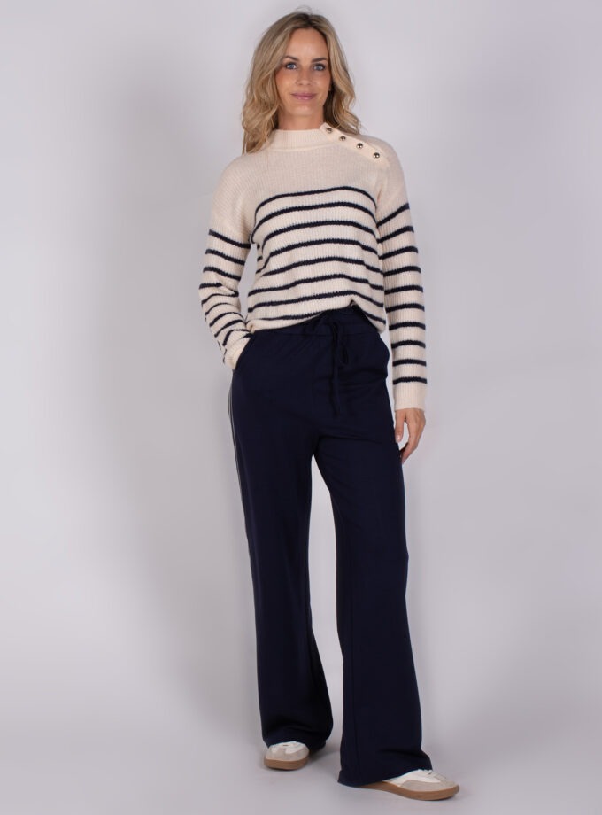 Vrouw draagt een gebreide trui met marineblauwe strepen en knopen op de schouder, gecombineerd met een donkerblauwe broek.