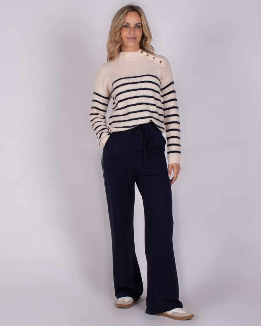 Vrouw draagt een gebreide trui met marineblauwe strepen en knopen op de schouder, gecombineerd met een donkerblauwe broek.