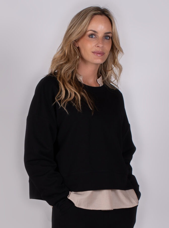 Vrouw poseert in een zwart sweatshirt met beige blouse eronder tegen een neutrale achtergrond.