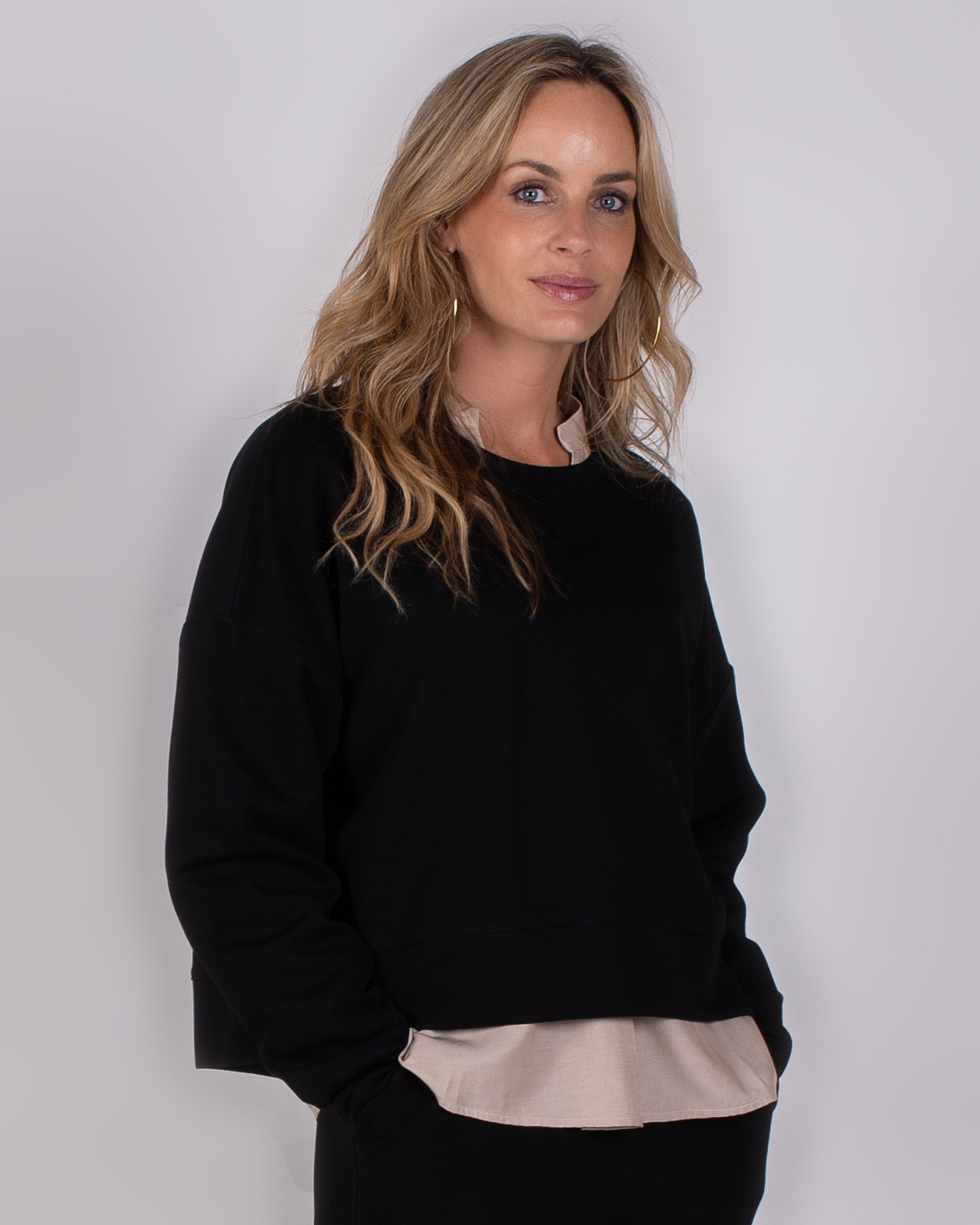 Vrouw poseert in een zwart sweatshirt met beige blouse eronder tegen een neutrale achtergrond.