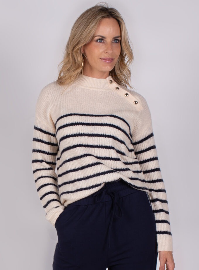 Vrouw draagt een gebreide trui met donkerblauwe strepen en knoopdetails op de schouder uit de Studio AW25-26 collectie.