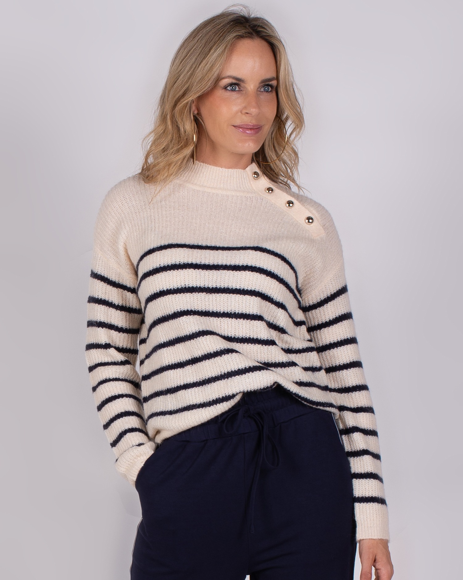 Vrouw draagt een gebreide trui met donkerblauwe strepen en knoopdetails op de schouder uit de Studio AW25-26 collectie.