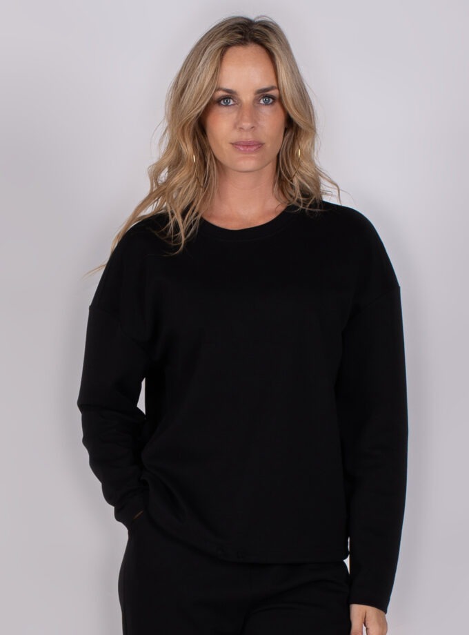 Vrouw poseert in een zwarte, lange mouwen trui uit de Studio AW25-26 collectie tegen een grijze achtergrond.