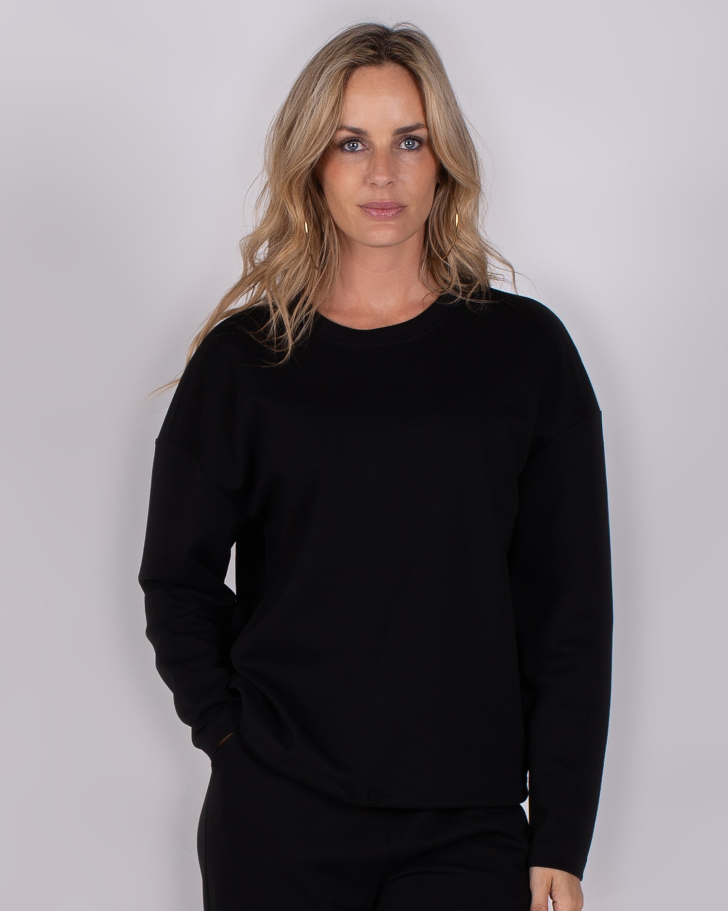 Vrouw poseert in een zwarte, lange mouwen trui uit de Studio AW25-26 collectie tegen een grijze achtergrond.
