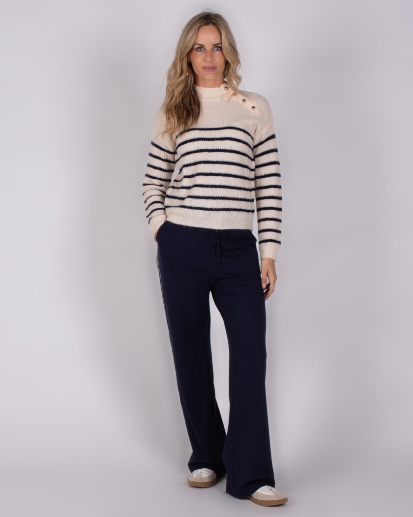 Vrouw draagt een crèmekleurige trui met donkerblauwe strepen en een donkerblauwe broek uit de Studio AW25-26 collectie.