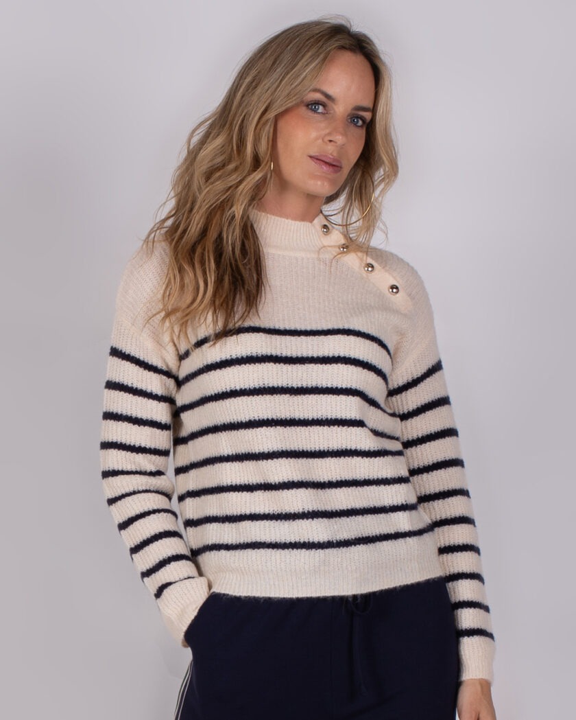 Vrouw draagt een crèmekleurige trui met donkerblauwe strepen en gouden knopen op de schouder uit de Studio AW25-26-collectie.