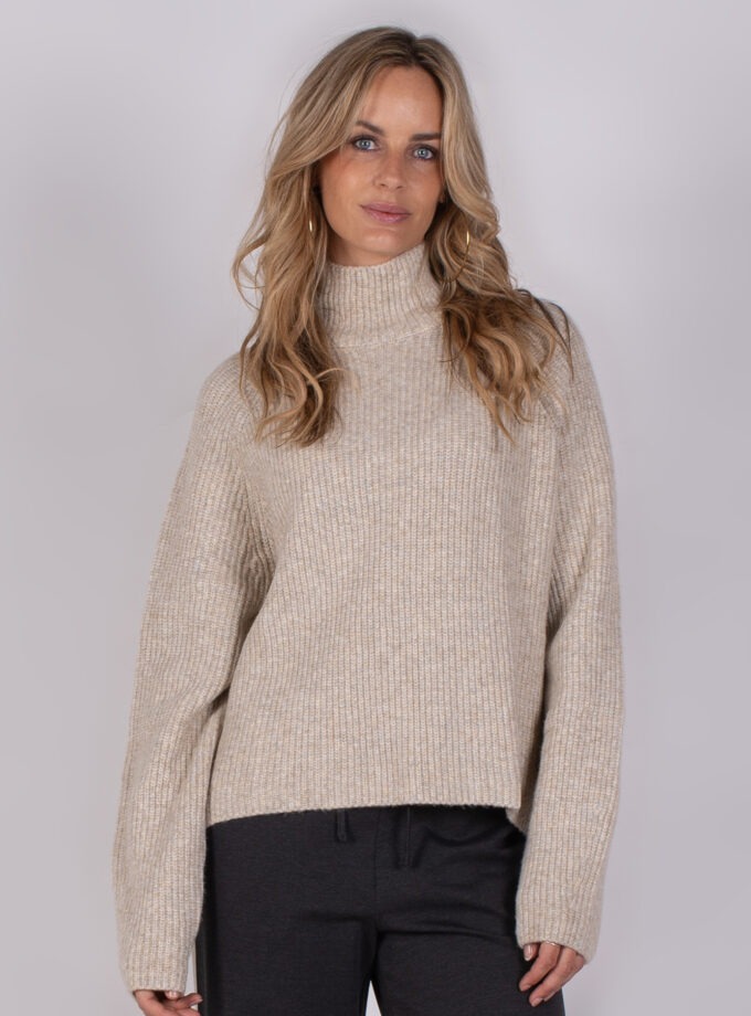 Vrouw draagt een beige, grofgebreide coltrui uit de Studio AW25-26 collectie.