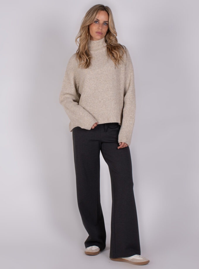 Vrouw draagt een beige gebreide coltrui en donkergrijze wijde broek uit de Studio AW25-26 collectie.