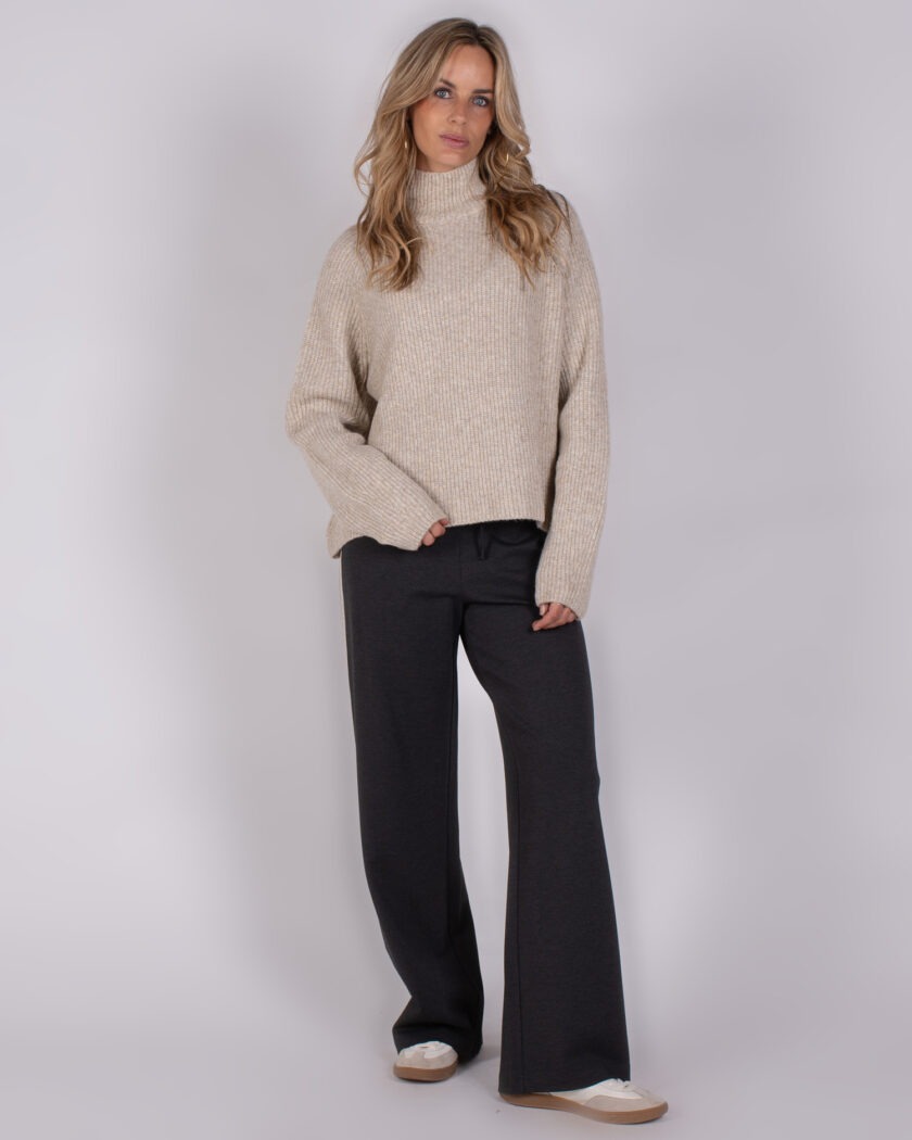 Vrouw draagt een beige gebreide coltrui en donkergrijze wijde broek uit de Studio AW25-26 collectie.