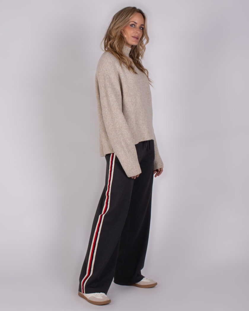 Model draagt een beige gebreide trui en zwarte broek met witte en rode zijstrepen uit de Studio AW25-26 collectie.