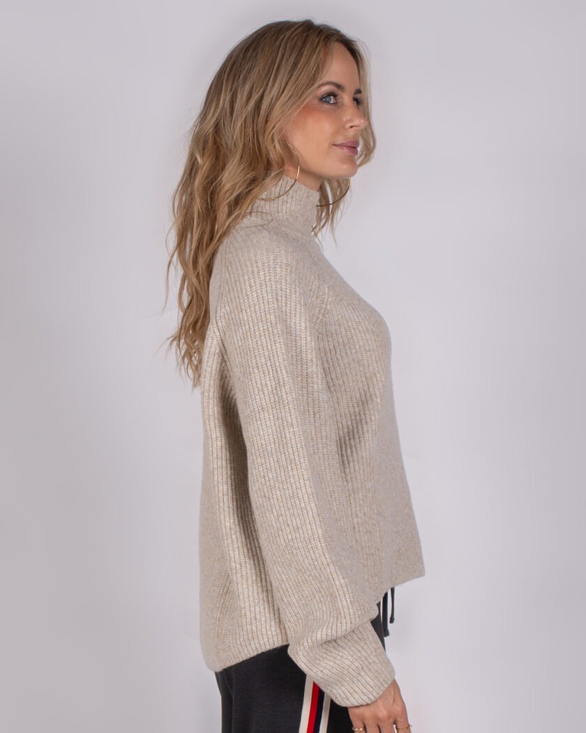 Vrouw in zijaanzicht draagt een beige, geribde coltrui uit de Studio AW25-26 collectie.