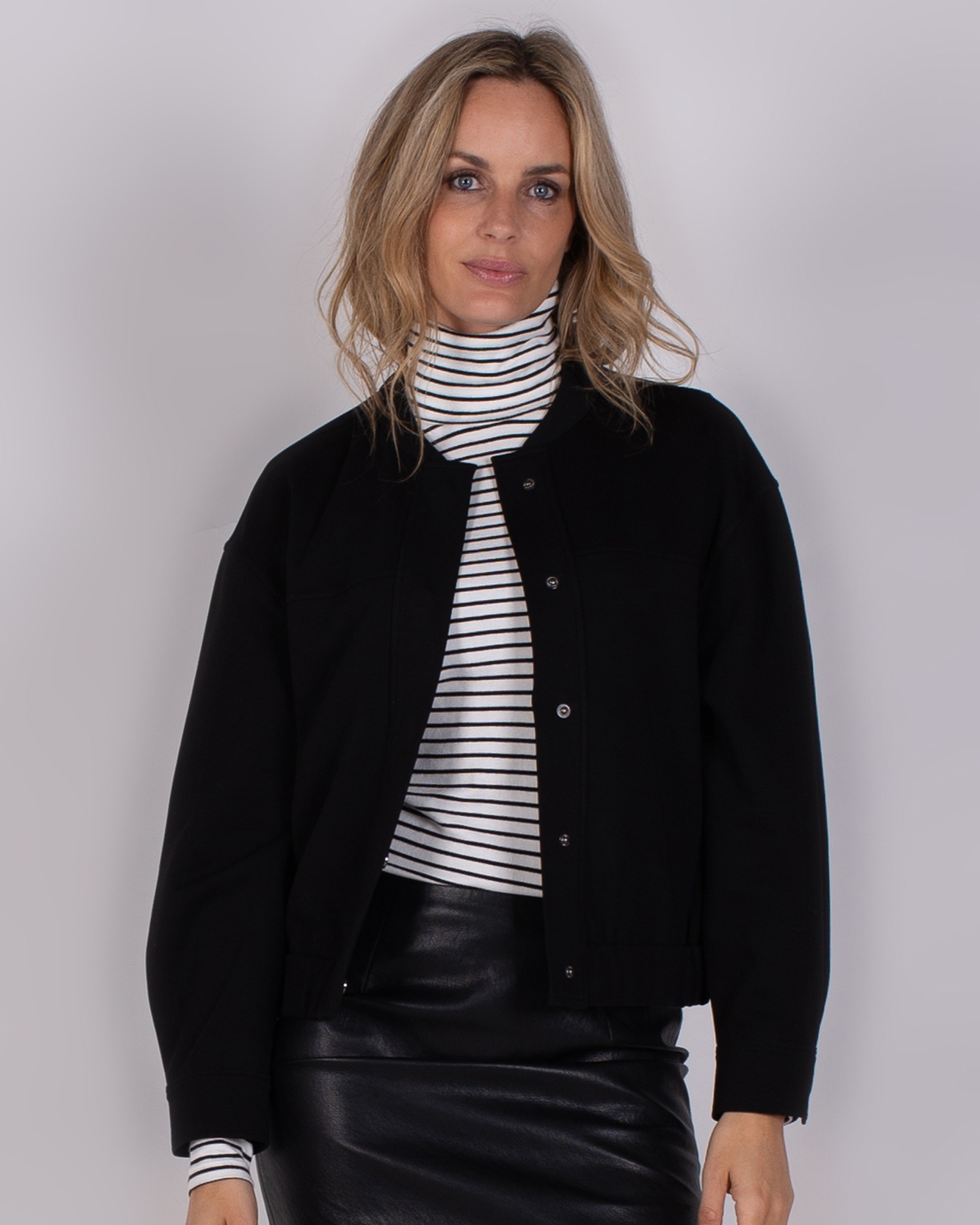 Vrouw draagt een zwart jack over een gestreepte coltrui en een zwarte leren rok uit de Studio AW25-26 collectie.