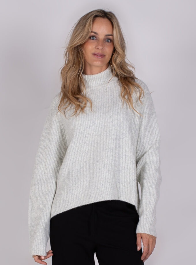 Vrouw draagt een lichtgrijze, geribde trui met col en zwarte broek uit de Studio AW25-26 collectie.