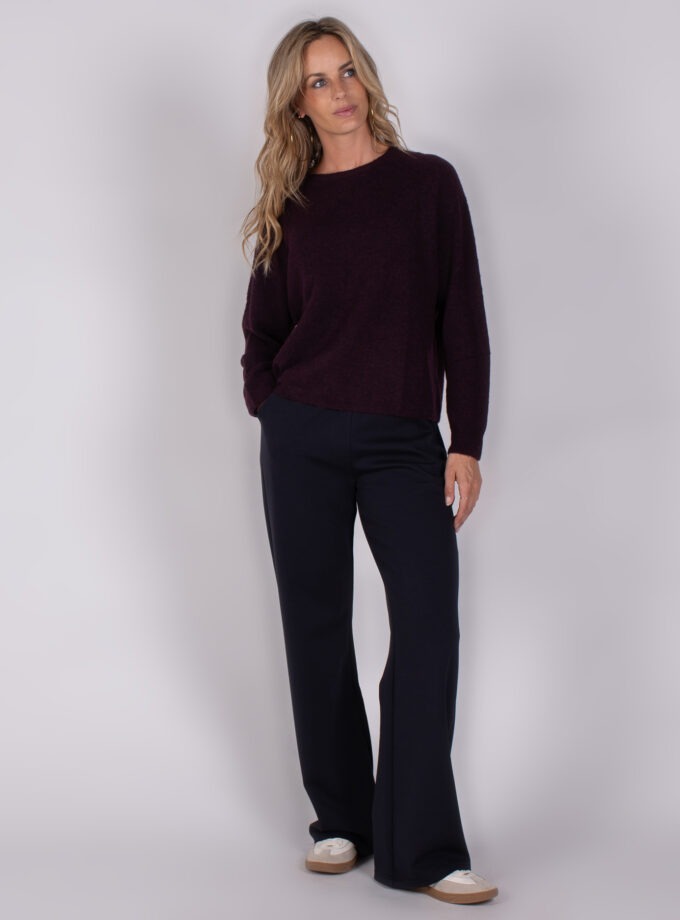 Vrouw draagt een donkerpaarse trui en wijde navy broek met witte sneakers uit de Studio AW25-26 collectie.