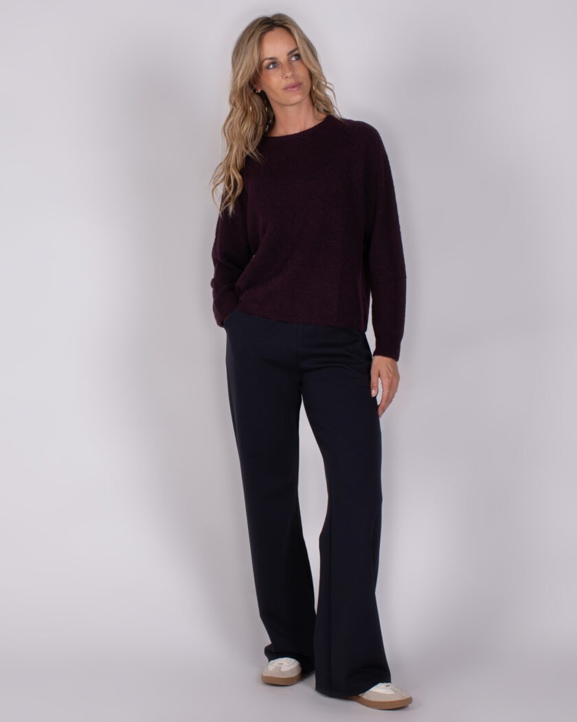 Vrouw draagt een donkerpaarse trui en wijde navy broek met witte sneakers uit de Studio AW25-26 collectie.