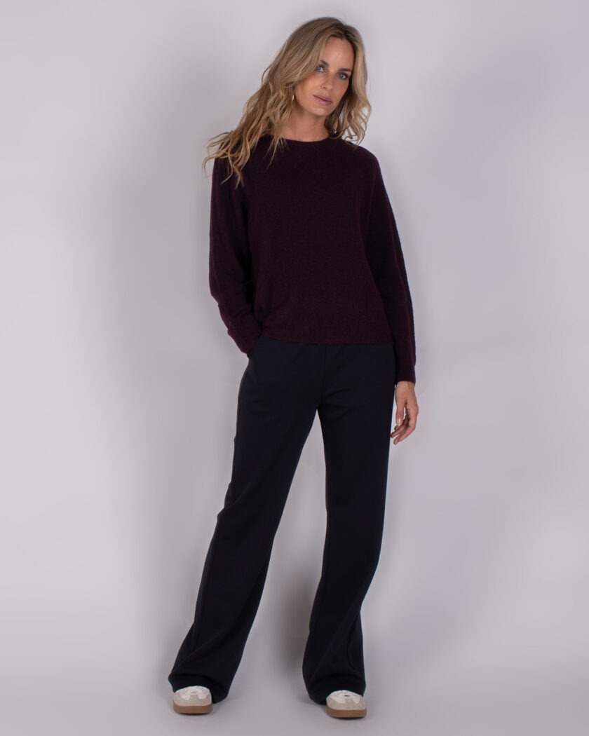 Vrouw draagt een donkerpaarse trui en donkerblauwe broek uit de Studio AW25-26 collectie.