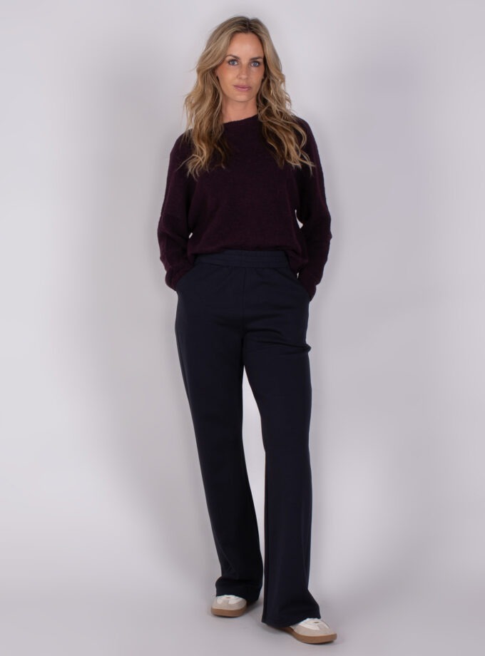 Vrouw draagt een donkerpaarse trui en donkerblauwe broek uit de Studio AW25-26 collectie.