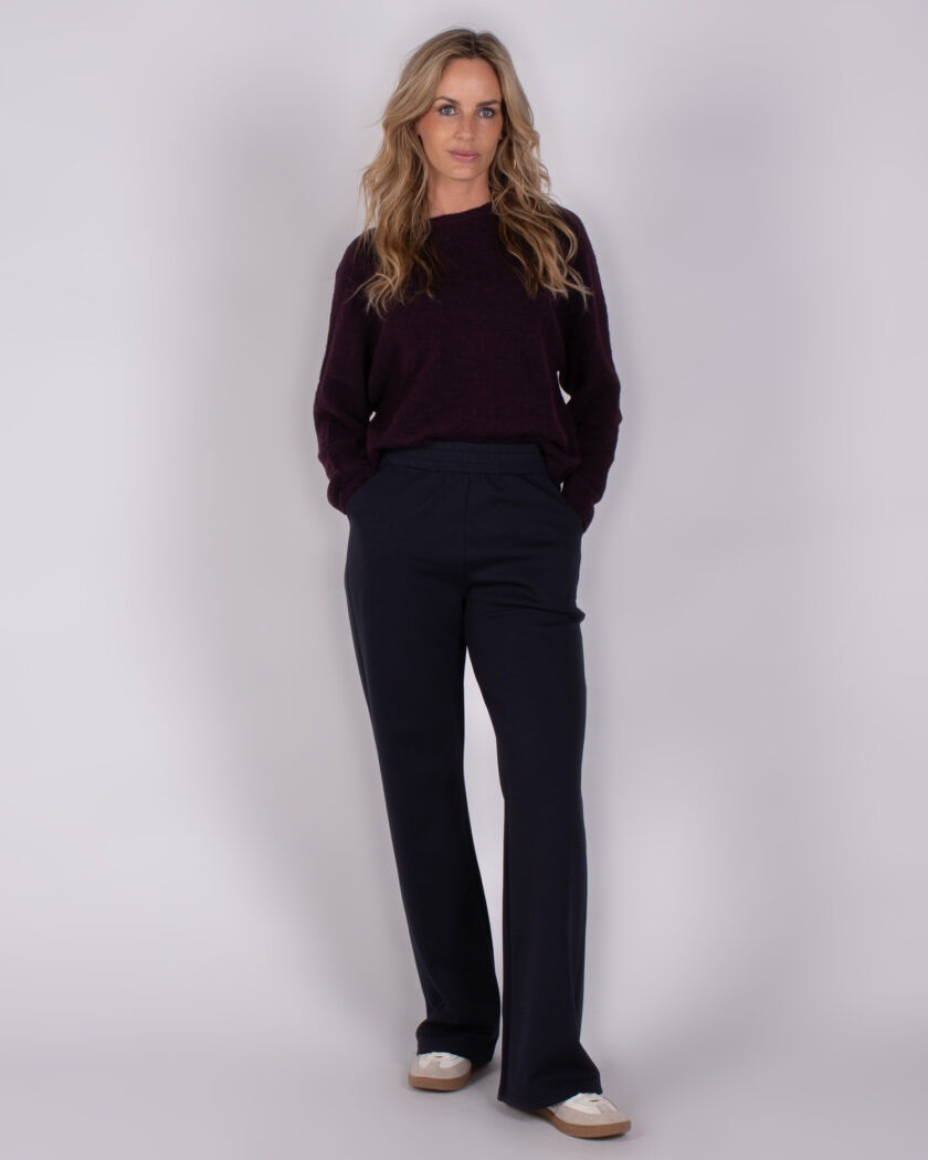 Vrouw draagt een donkerpaarse trui en donkerblauwe broek uit de Studio AW25-26 collectie.