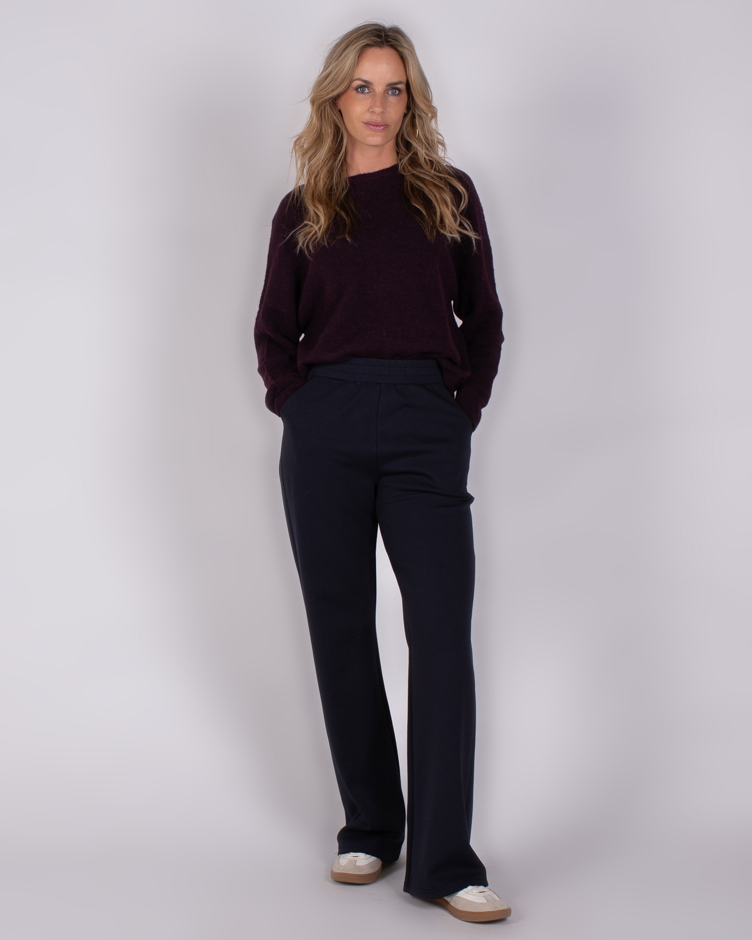 Vrouw draagt een donkerpaarse trui en donkerblauwe broek uit de Studio AW25-26 collectie.