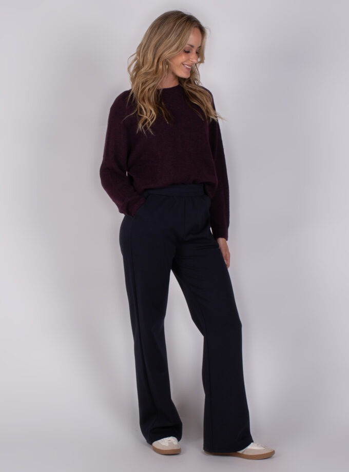 Vrouw draagt een donkerpaarse trui en donkerblauwe wijde broek uit de Studio AW25-26 collectie.