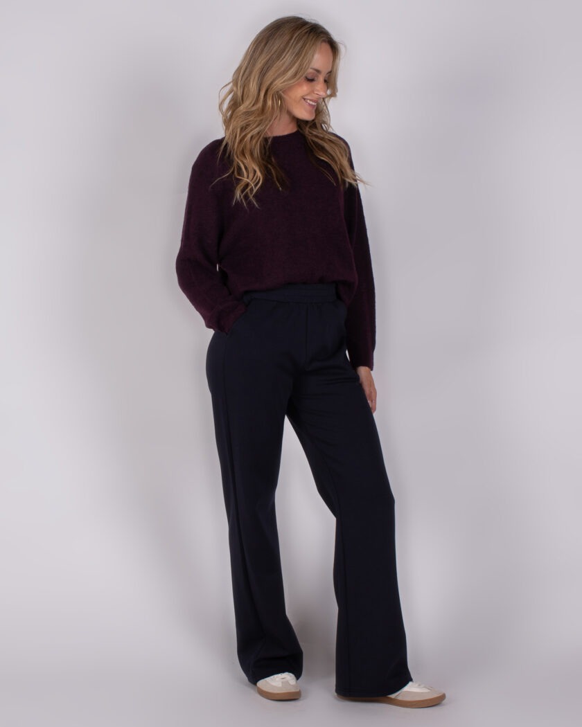 Vrouw draagt een donkerpaarse trui en donkerblauwe wijde broek uit de Studio AW25-26 collectie.