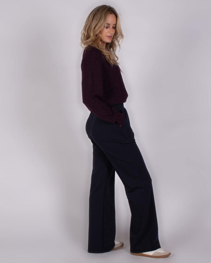 Vrouw in zijaanzicht draagt een donkerpaarse trui en wijde navy broek uit de Studio AW25-26 collectie.