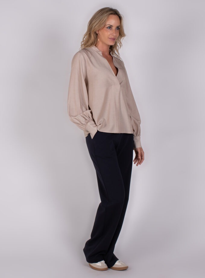 Vrouw draagt een beige blouse met lange mouwen en zwarte broek uit de Studio AW25-26 collectie.
