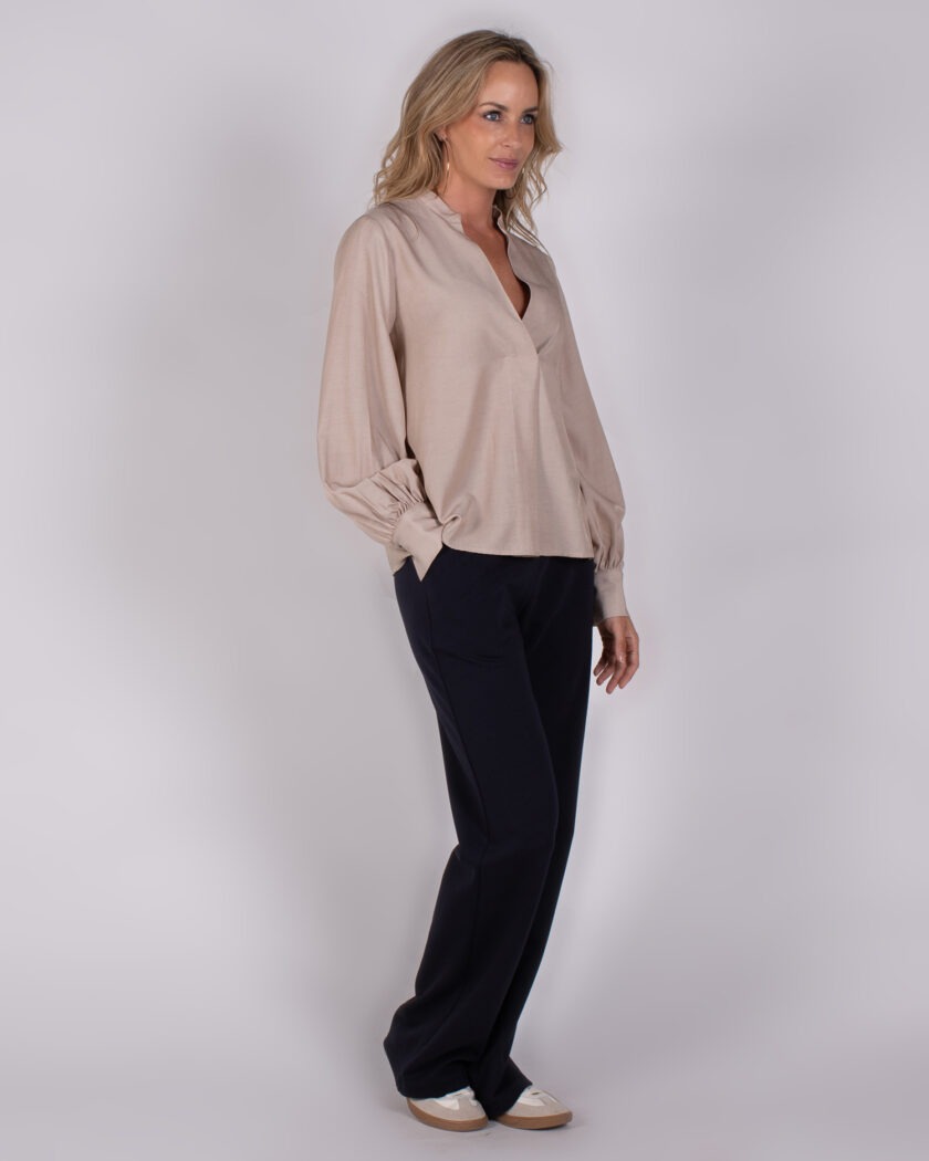 Vrouw draagt een beige blouse met lange mouwen en zwarte broek uit de Studio AW25-26 collectie.