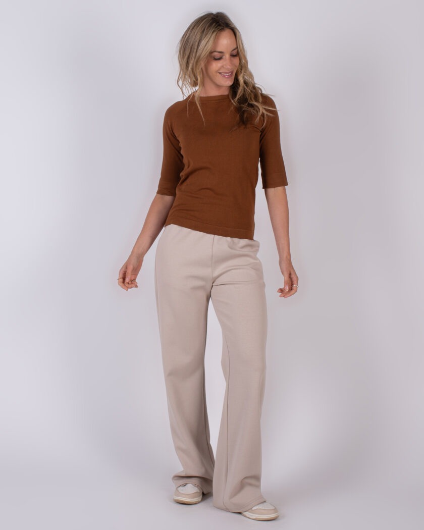 Vrouw in een bruine top met halve mouwen en beige, wijdvallende broek poseert tegen een neutrale achtergrond.