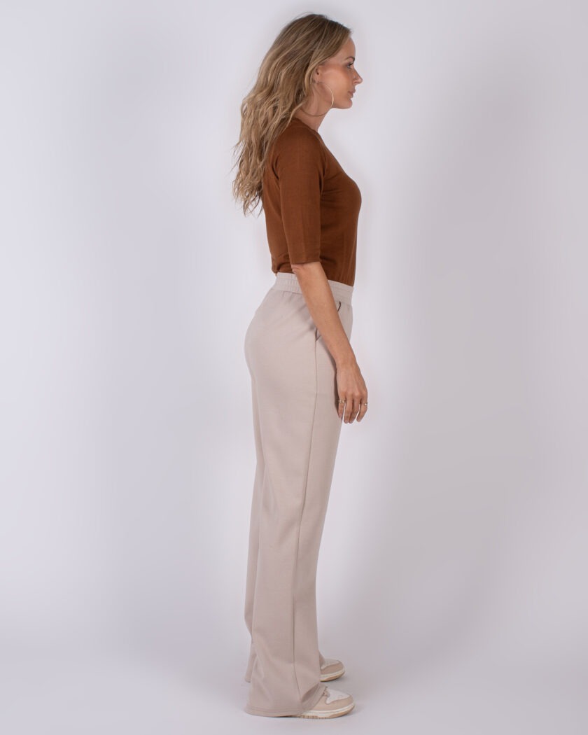 Vrouw in zijaanzicht draagt een bruin shirt met beige, wijde broek en sneakers op een neutrale achtergrond.