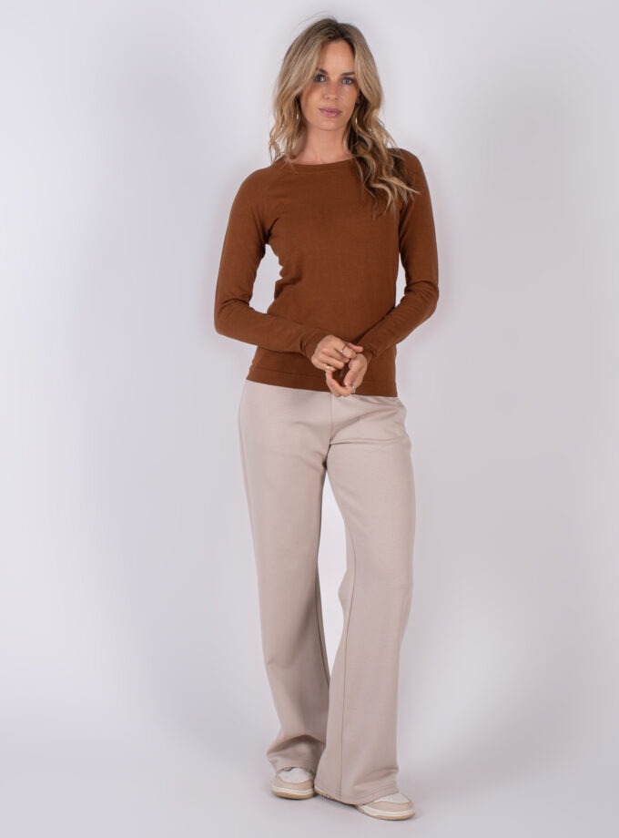 Vrouw in een bruine trui en beige, wijde broek poseert in studio tegen een neutrale achtergrond.