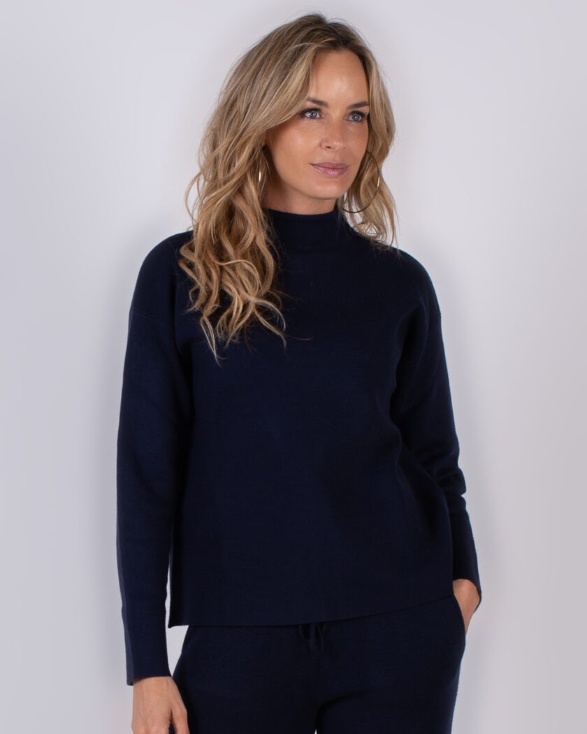 Vrouw draagt een donkerblauwe trui met hoge kraag en bijpassende broek uit de Studio AW25-26 collectie.