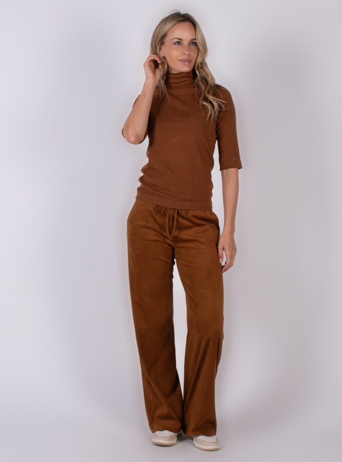 Vrouw in een bruin Studio AW25-26 outfit met coltrui en losse broek poseert tegen een neutrale achtergrond.
