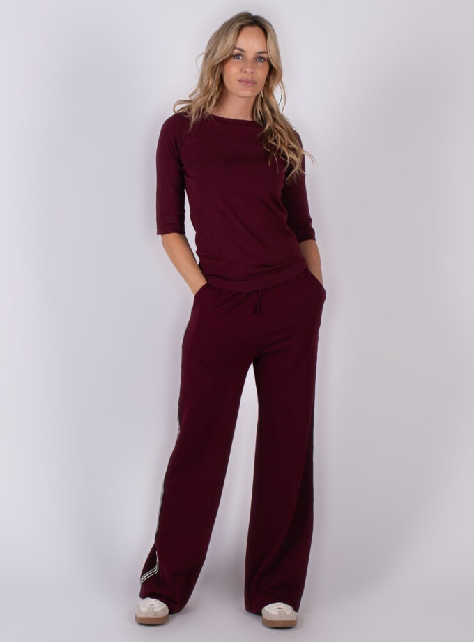 Vrouw poseert in een bordeauxrood loungepak met wijde broek en halve mouw, uit de Studio AW25-26 collectie.