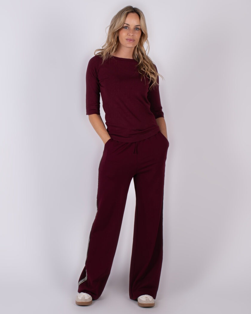 Vrouw poseert in een bordeauxrood loungepak met wijde broek en halve mouw, uit de Studio AW25-26 collectie.