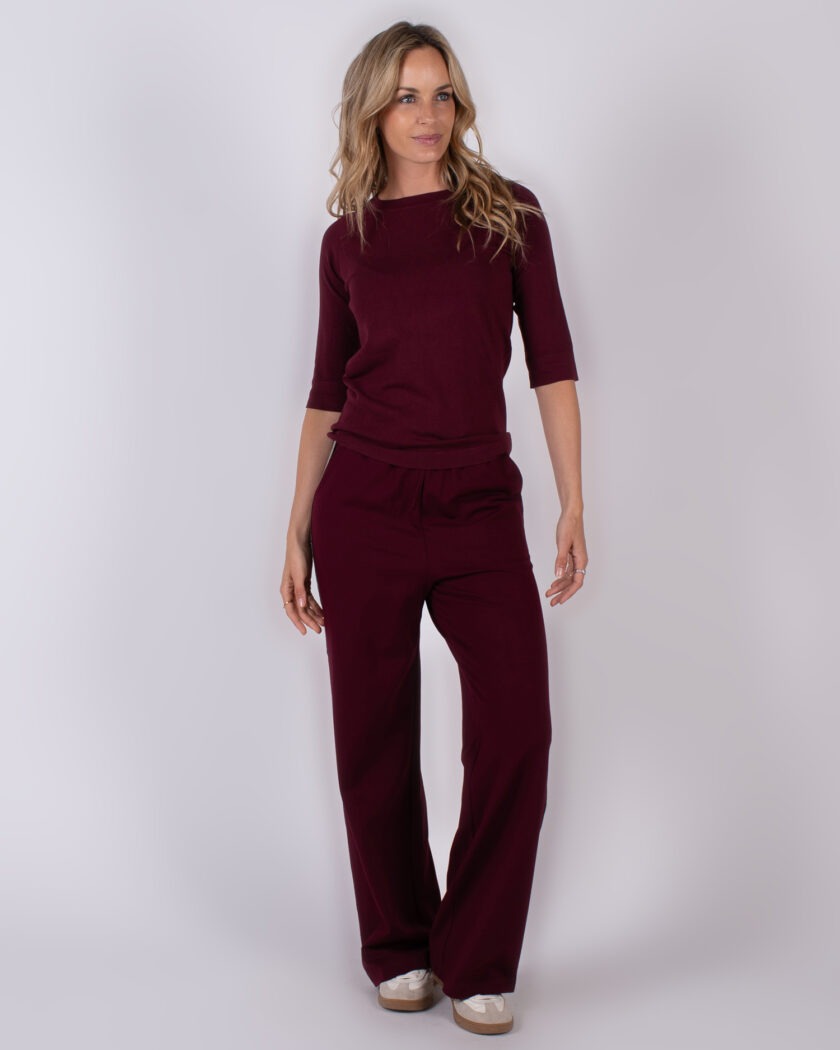 Vrouw draagt een bordeauxrode lounge-outfit met wijde broek en halfmouw top uit Studio AW25-26 collectie.