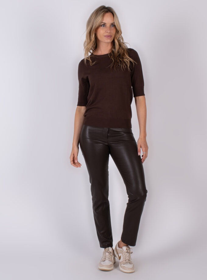 Vrouw draagt een donkerbruine top met halve mouwen, een donkerbruine leren broek en beige sneakers tegen een neutrale achtergrond.