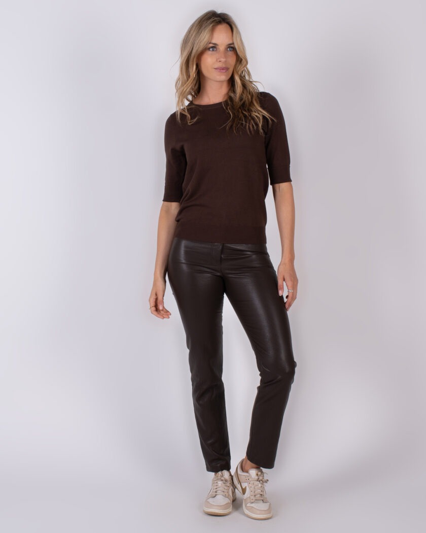 Vrouw draagt een donkerbruine top met halve mouwen, een donkerbruine leren broek en beige sneakers tegen een neutrale achtergrond.