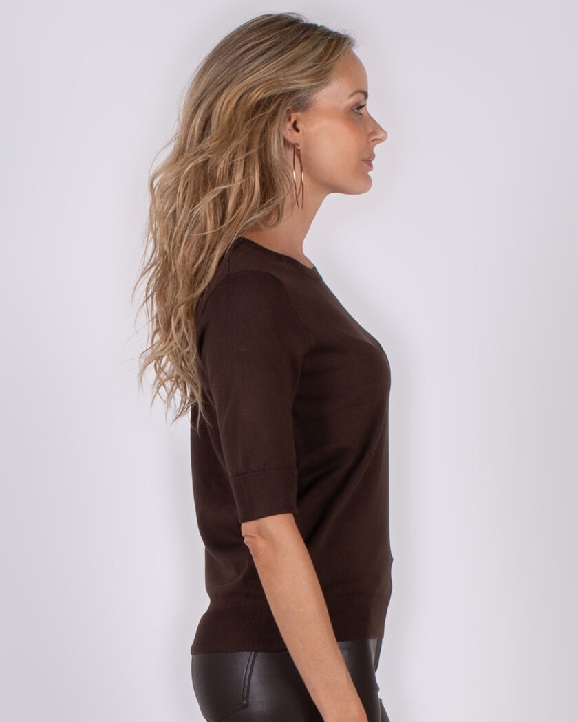 Vrouw in profiel poseert in een donkerbruin shirt met korte mouwen en zwarte leren broek tegen een neutrale achtergrond.