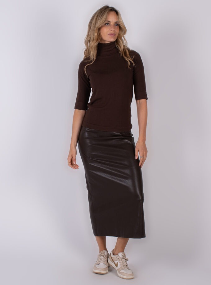 Vrouw draagt een donkerbruine coltrui met half lange mouwen en een glanzende, donkerbruine leren midi-rok, gecombineerd met beige sneakers.