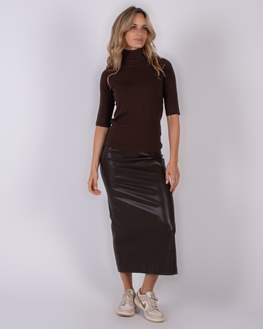 Vrouw draagt een donkerbruine coltrui met half lange mouwen en een glanzende, donkerbruine leren midi-rok, gecombineerd met beige sneakers.